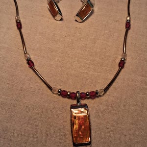 Puede incluir: Un conjunto de joyería compuesto por un collar y pendientes a juego. El collar presenta un colgante rectangular con una piedra de color ámbar, acentuado por cuentas de color rojo oscuro y tubos de metal plateado. Los pendientes reflejan el diseño del colgante.
