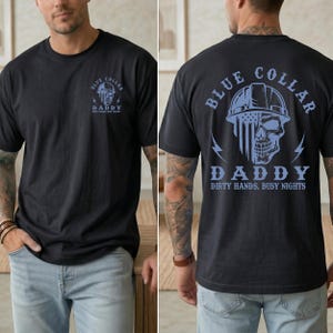 May include: A black t-shirt featuring a light blue graphic of a skull wearing a hard hat with a flag pattern. The front has a small chest logo, while the back displays the same design with the text "BLUE COLLAR DADDY DIRTY HANDS, BUSY NIGHTS."