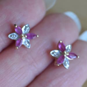 May include: A pair of flower-shaped stud earrings. Each earring features a gold-colored base with six petals. The petals alternate between white and magenta-colored stones, creating a floral design. The earrings are small and delicate.