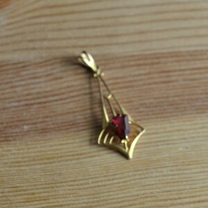 May include: A gold pendant with a red gemstone in the center. The pendant is shaped like a teardrop and has a delicate design.