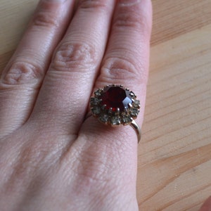 May include: A ring with a large red gemstone surrounded by smaller clear gemstones.