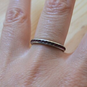 May include: A silver ring with a band of small, dark red gemstones. The ring is on a person's finger, with the hand in the foreground. The ring's gemstones are set closely together, creating a continuous line of color.