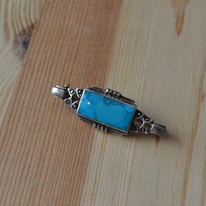 May include: A silver brooch with a turquoise stone set in a rectangular shape. The brooch has a delicate design with intricate details.