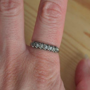 May include: A silver-toned band ring featuring a row of five small, round, clear stones set in individual circular metal settings. The ring is worn on a finger, showcasing the metallic finish and the alignment of the stones across the band.