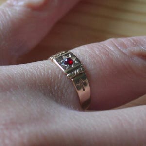 May include: A gold ring featuring a central square setting with a star-shaped engraving and a small, round, deep red gemstone. The band includes a twisted metal detail near the setting and a fleur-de-lis symbol engraved on the side of the shank.