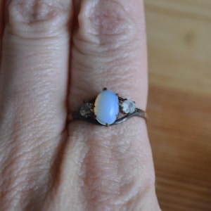 May include: A silver ring with an oval, light blue gemstone as the centerpiece, flanked by two smaller, round, clear stones. The ring is set against a person's skin, with the focus on the jewelry.