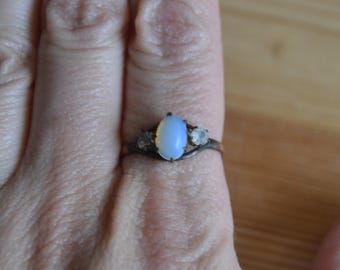 May include: A silver ring with an oval, light blue gemstone as the centerpiece, flanked by two smaller, round, clear stones. The ring is set against a person's skin, with the focus on the jewelry.