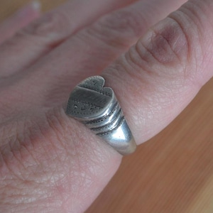 May include: A silver-toned ring featuring a heart-shaped face with engraved decorative patterns. The band of the ring displays a series of three horizontal ridges that transition into a smooth, solid shank, encircling the finger with a metallic finish.