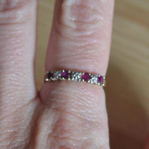 May include: A gold band ring set with alternating red gemstones and clear stones. The ring is a classic design, featuring a row of vibrant red stones and sparkling clear stones. The ring is shown on a finger.