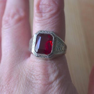 May include: A silver-toned ring featuring a large, rectangular, faceted deep red gemstone set in a bezel. The metal band displays an intricate engraved leaf pattern on the sides and a textured border surrounding the central red stone.