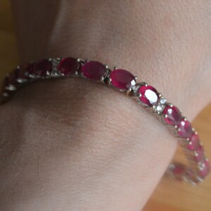 May include: A silver-toned tennis bracelet featuring a series of oval-cut, deep red rubies set in metal links. Each ruby is separated by small, clear, round-cut stones, creating a continuous, repeating pattern around the entire length of the bracelet.