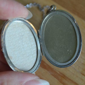 May include: Open silver oval locket on a silver chain. The locket is open, revealing two oval spaces. One side has a light beige paper insert, and the other side is a reflective silver color. The locket is held by a hand.