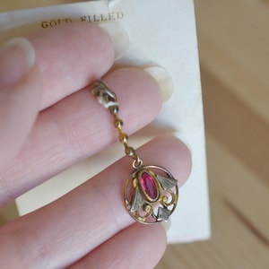 May include: A gold-filled earring with a red gemstone center. The earring has a decorative circular design with silver accents and a delicate chain. The text "GOLD FILLED" is visible on the attached card.
