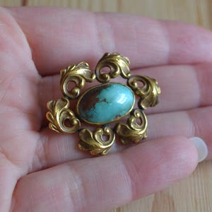 May include: A vintage-style brooch featuring a central oval turquoise stone with brown inclusions, set in a gold-toned metal frame. The frame has ornate, swirling leaf-like designs around the stone, creating a floral pattern.