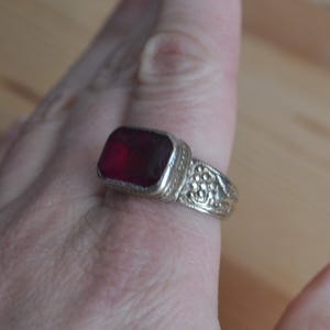 May include: A silver ring with a large, rectangular, deep red gemstone. The ring has an ornate, detailed design around the gemstone and on the band. The ring is worn on a finger.