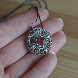 May include: A silver pendant necklace with a red gemstone in the center. The pendant is circular and has a delicate, intricate design.