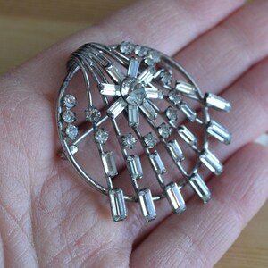 May include: A silver brooch with a geometric design featuring a central starburst of clear rhinestones surrounded by smaller rhinestones.