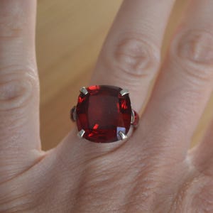 May include: A silver ring featuring a large, square-cut, deep red gemstone. The gemstone is set with four prongs and has a brilliant, reflective surface. The ring is displayed on a person's hand, showcasing the jewelry's design and color.