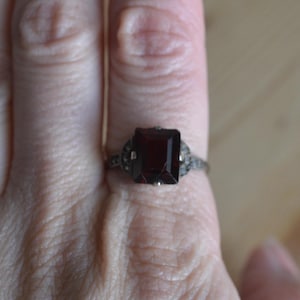 May include: A silver ring featuring a large, rectangular, deep red gemstone. The ring has a decorative setting with smaller accent stones on either side of the main stone. The ring is shown on a finger.