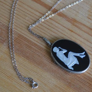 May include: Silver-toned necklace with an oval pendant. The pendant features a black background with a white silhouette of a reclining figure. The chain is delicate and the clasp is visible.