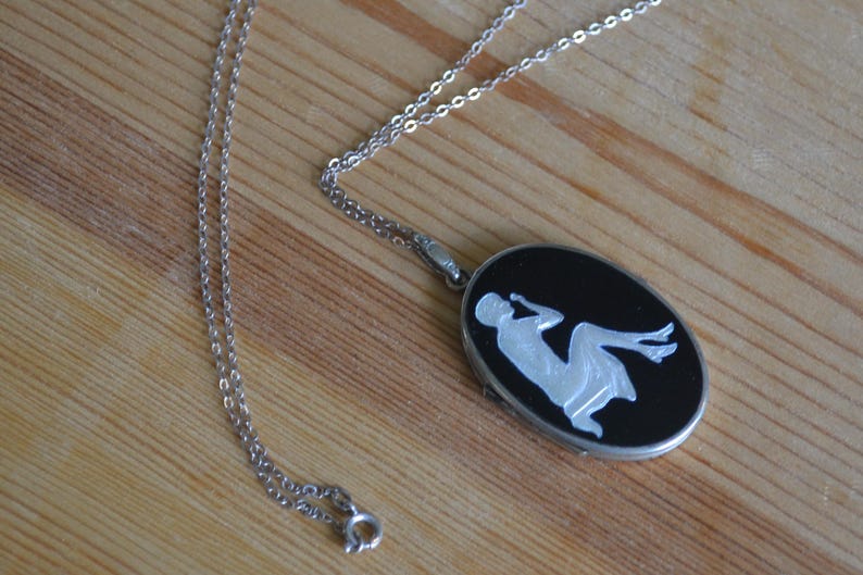 May include: Silver-toned necklace with an oval pendant. The pendant features a black background with a white silhouette of a reclining figure. The chain is delicate and the clasp is visible.