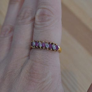 May include: A gold ring featuring alternating oval-cut, deep red gemstones and small, round, clear gemstones. The ring is set on a gold band and is displayed on a person's finger.