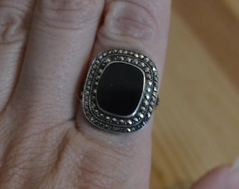 May include: Silver-toned ring with a square, black onyx center stone. The ring is surrounded by small, round, silver-colored accents. The ring is on a finger, with a blurred background.