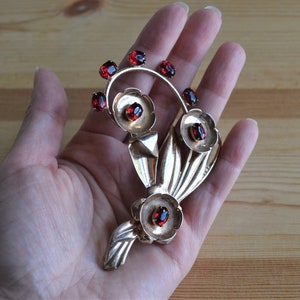 May include: A silver brooch with a floral design featuring red gemstones. The brooch is shaped like a bouquet of flowers with leaves and stems.