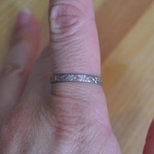 May include: A silver ring with a repeating floral pattern. The ring is worn on a finger, showcasing the intricate design. The ring's surface reflects light, highlighting the details of the engraved flowers and leaves. A piece of jewelry.