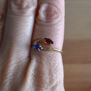 May include: A gold-colored ring with a bypass design, featuring a blue and a red gemstone. The gemstones are oval-shaped and set in a delicate, curved band. The ring is shown on a finger.