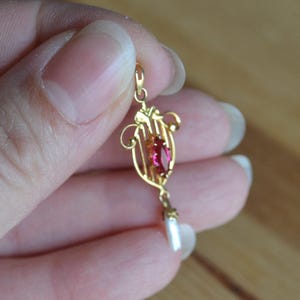 May include: A gold-colored pendant with a red gemstone and a white pearl drop. The pendant has an ornate, filigree design with a central, oval-shaped red stone. A small, white pearl hangs from the bottom.