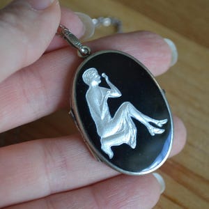 May include: Silver-toned oval locket with a black enamel background. The locket features a detailed, seated figure in a flowing gown. The figure is rendered in silver against the black enamel. The locket is suspended from a delicate chain.