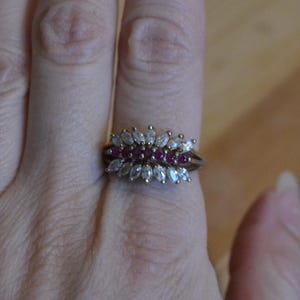 May include: A gold-toned ring featuring a cluster of gemstones. The ring has a central row of small, round, red stones, flanked by rows of marquise-cut, clear stones. The ring is worn on a finger.