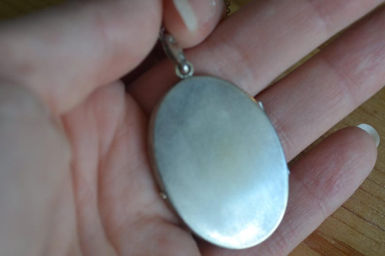 May include: Silver-toned oval locket pendant. The locket is held in a hand, showcasing its smooth, reflective surface. The pendant hangs from a delicate chain, suggesting a piece of jewelry. The locket's design is simple and elegant.