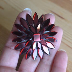 May include: A decorative, flower-shaped brooch with a central square and petal-like shapes. The brooch is made of reflective, ruby-red materials, creating a sparkling effect. The brooch is held in a hand.