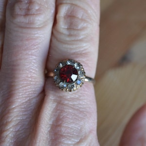 May include: A gold ring featuring a large, faceted, deep red gemstone at the center, surrounded by smaller, clear and blue stones. The ring is set in a gold band, creating a classic and elegant design. The ring is shown on a finger.