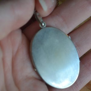 May include: Silver-toned oval locket pendant. The locket is held in a hand, showcasing its smooth, reflective surface. The pendant hangs from a delicate chain, suggesting a piece of jewelry. The locket's design is simple and elegant.