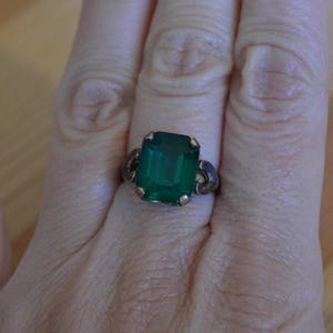 May include: A vintage ring featuring a large, emerald-cut green gemstone. The ring has a dark, possibly antique, metal band with decorative side details. The gemstone is a vibrant green color, set in a gold-toned bezel.