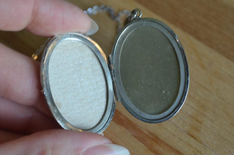 May include: Open silver oval locket on a silver chain. The locket is open, revealing two oval spaces. One side has a light beige paper insert, and the other side is a reflective silver color. The locket is held by a hand.