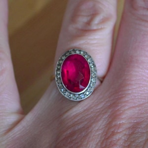 May include: A close-up of a ring featuring a large, oval-shaped, vibrant red gemstone. The gemstone is surrounded by a halo of smaller, clear stones. The ring's band appears to be gold-colored, creating a striking contrast with the red and clear stones.