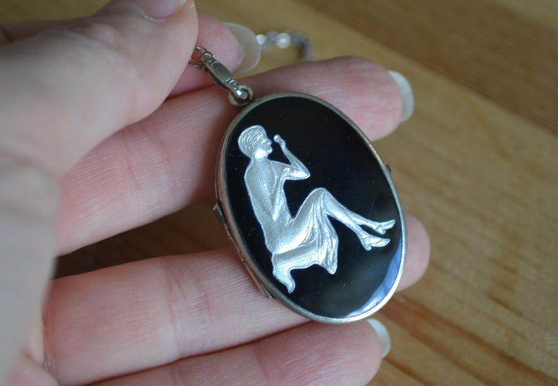 May include: Silver-toned oval locket with a black enamel background. The locket features a detailed, seated figure in a flowing gown. The figure is rendered in silver against the black enamel. The locket is suspended from a delicate chain.