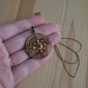 May include: A gold pendant necklace with a round, filigree design and a red gemstone in the center. The pendant is hanging from a gold chain.