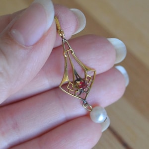 May include: A gold-toned earring with an Art Deco design, featuring a red gemstone and a dangling pearl. The earring is held in a hand, showcasing its intricate filigree and elegant style. The background is a blurred wooden surface.
