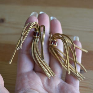 May include: A pair of gold-toned tassel earrings with a rectangular, dark red gemstone accent. The earrings are held in a hand against a blurred wooden background. The tassels are made of thin, woven strands.