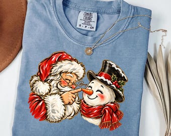 Könnte beinhalten: Ein hellblaues T-Shirt mit einer Grafik vom Weihnachtsmann, der die orangefarbene Karottennase eines Schneemanns berührt. Der Schneemann trägt einen schwarzen Zylinder mit Stechpalme und einen roten Schal. Der Text Boop steht dazwischen.