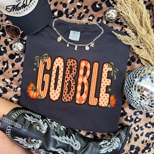 K&ouml;nnte beinhalten: Ein dunkelgraues Kurzarm-T-Shirt mit dem Wort GOBBLE in gro&szlig;en, gemusterten orangefarbenen und beigen Buchstaben. Das Design umfasst Punkte, Streifen, Rautenmuster und Leopardenmuster, akzentuiert durch kleine gr&uuml;ne Schleifen, Sterne und einen K&uuml;rbis.