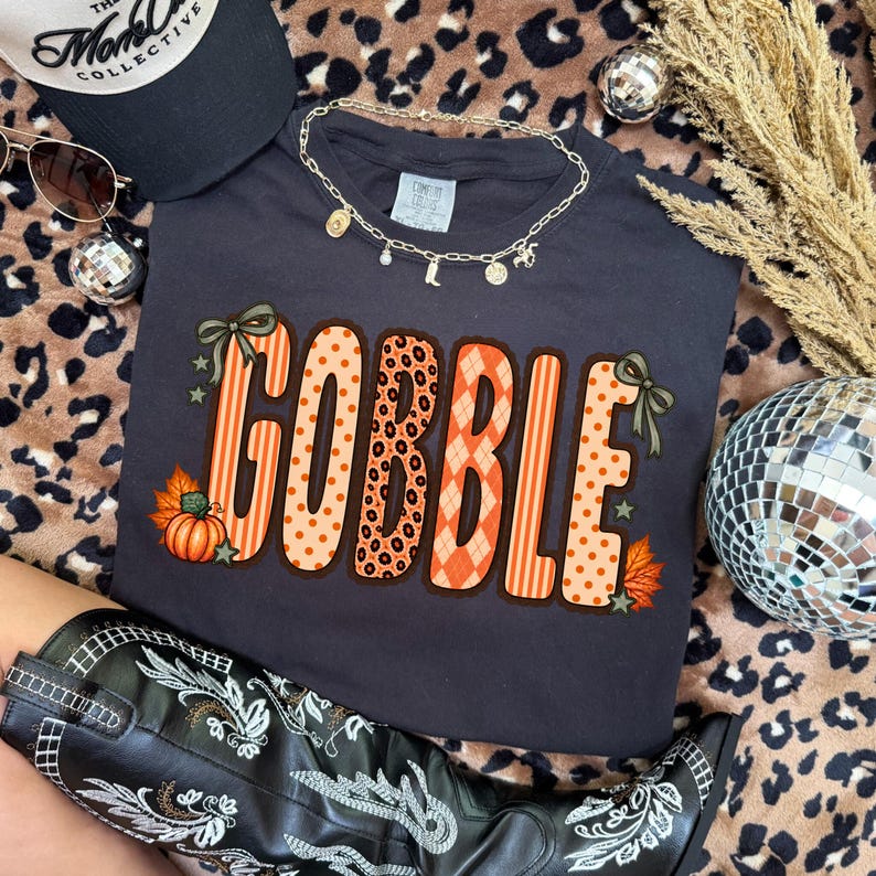 K&ouml;nnte beinhalten: Ein dunkelgraues Kurzarm-T-Shirt mit dem Wort GOBBLE in gro&szlig;en, gemusterten orangefarbenen und beigen Buchstaben. Das Design umfasst Punkte, Streifen, Rautenmuster und Leopardenmuster, akzentuiert durch kleine gr&uuml;ne Schleifen, Sterne und einen K&uuml;rbis.