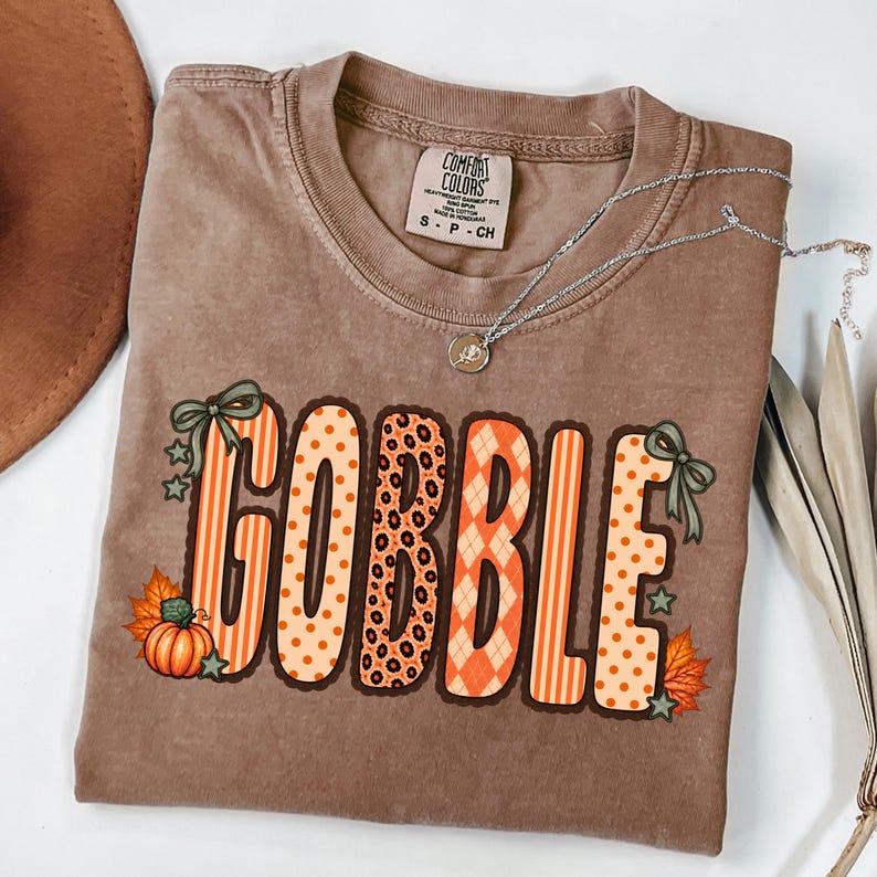 K&ouml;nnte beinhalten: Ein braunes Kurzarm-T-Shirt mit einem Comfort Colors-Etikett am Halsausschnitt. Auf der Vorderseite steht das Wort GOBBLE in gro&szlig;en, gemusterten orangefarbenen und beigen Buchstaben, verziert mit gr&uuml;nen Schleifen, K&uuml;rbissen und Herbstbl&auml;ttern.