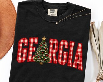Könnte beinhalten: Ein schwarzes kurzärmeliges T-Shirt mit einem Comfort Colors-Etikett am Halsausschnitt. Auf der Vorderseite steht das Wort GEORGIA in rot-weißem Vichy-Karo, wobei ein geschmückter Weihnachtsbaum den Buchstaben O ersetzt.