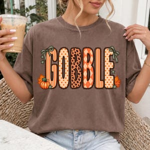 K&ouml;nnte beinhalten: Ein braunes Kurzarm-T-Shirt mit dem Wort GOBBLE in gro&szlig;en, orangefarbenen Buchstaben, die verschiedene Muster wie Streifen, Punkte, Leopardenmuster und Karos aufweisen, verziert mit kleinen gr&uuml;nen Schleifen, Sternen und Herbstbl&auml;ttern.